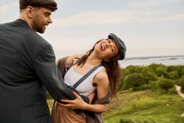 Smiling and bearded man in jacket and newsboy cap hugging cheerful girlfriend in vest and suspenders while standing with nature and sky at background, fashion-forwards in countryside clipart