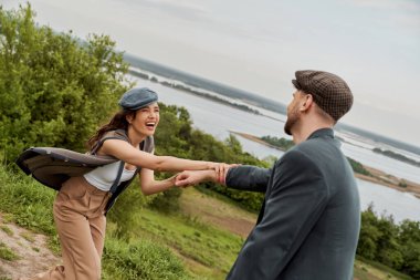 Fashionable and cheerful brunette woman in vest and newsboy cap holding hand of bearded boyfriend in jacket while standing with nature at background, fashion-forwards in countryside clipart