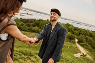 Tarz sahibi gazeteci çocuk şapkası ve ceketiyle gülümseyen ve sakallı adam bulanık esmer kız arkadaşının elini tutarken arka planda bulanık doğa ve ahşap, kırsalda moda ileri görüşlü