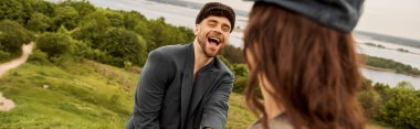 Excited and fashionable bearded man in newsboy cap and jacket standing near blurred brunette girlfriend with scenic nature at background, fashion-forwards in countryside, banner  clipart