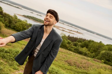 Şık giyinmiş neşeli sakallı adam ve gazeteci çocuk şapkası kız arkadaşının elini tutarken arka planda bulanık gökyüzü, kırsal kesimde moda ileri görüşlü.