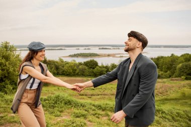 Positive and fashionable brunette woman in newsboy cap and vest holding hand of bearded boyfriend in jacket while standing with scenic landscape at background, fashionable couple in countryside clipart