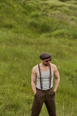 Eski moda gözlüklü, güneş gözlüklü, pantolon cebinde el ele tutuşan, arka planda bulanık çimenli çayırlarda dikilen, kır hayatının tadını çıkaran şık bir adam.
