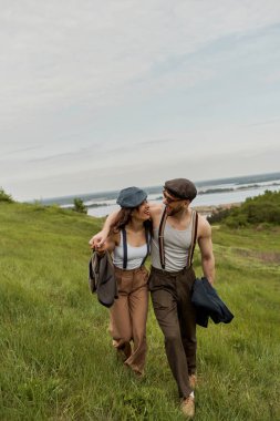 Neşeli ve modaya uygun romantik bir çift, gazeteci çocuk şapkaları, klasik kıyafetler ve pantolon askıları arka planda çimenli tepede sarılıp yürüyorlar, kırsal alanda moda bir çift.