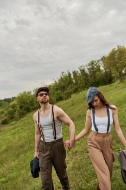 Havalı sakallı, güneş gözlüklü, pantolon askısı takan, esmer kız arkadaşı olan, gazeteci şapkalı, çimenli çimlerde yürüyen, kırsal alanda moda bir çift.