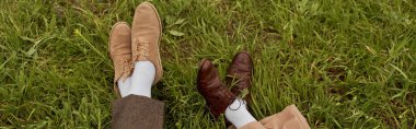 Top view of legs of romantic couple in pants and vintage shoes sitting together on green grassy meadow, stylish partners in rural escape, romantic getaway, banner clipart