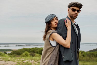 Gazeteci şapkalı ve yelekli esmer kadının portresi güneş gözlüklü ve ceketli sakallı erkek arkadaşı kucaklarken arka planda bulanık manzara, kırsal alanda moda ikilisi