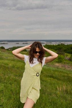 Trendy and smiling brunette woman in sunglasses and stylish sundress touching head and standing while spending time on blurred grassy meadow at background, summertime joy clipart