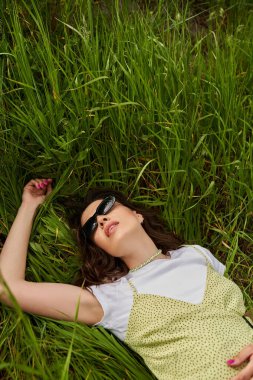 Yazın yeşil çimlerin üzerinde uzanan güneş gözlüklü, şık esmer kadın manzarası doğal manzara ve kırsal alanda rahatlama.