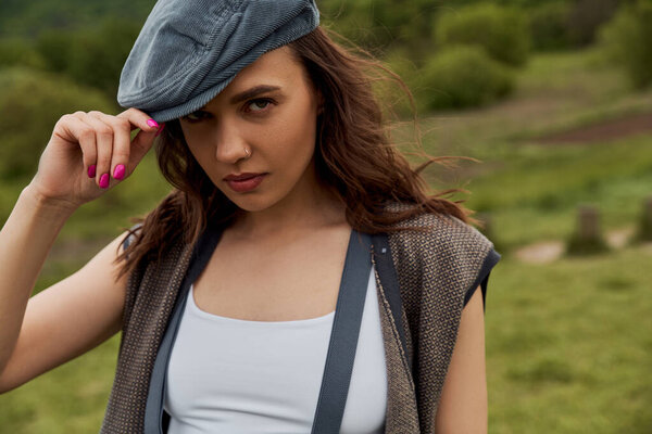 Portrait of stylish brunette woman in vintage clothes, vest and suspenders touching newsboy cap and looking at camera with blurred landscape at background, fashion-forward in countryside
