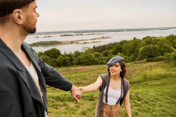 Smiling and trendy brunette woman in suspenders and newsboy cap holding hand of blurred boyfriend in jacket while standing with nature and sky at background, fashionable couple in countryside