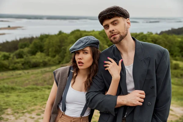 stock image Fashionable brunette woman in suspenders and newsboy cap hugging bearded boyfriend in jacket and looking at camera together with blurred landscape at background, fashionable couple in countryside