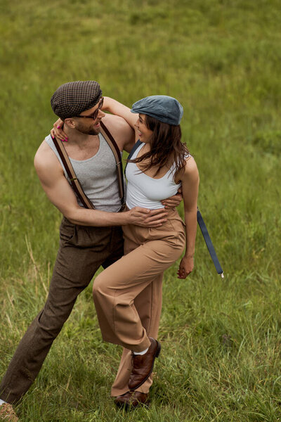 Positive and trendy bearded man in sunglasses hugging brunette girlfriend in newsboy cap and suspenders while standing on grassy field at background, stylish pair amidst nature
