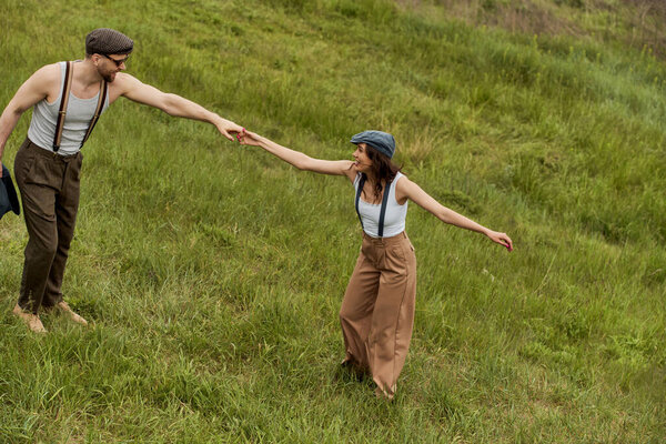 Side view of positive and fashionable couple in newsboy caps and susenders holding hands and having fun on grassy meadow at background, stylish couple enjoying country life