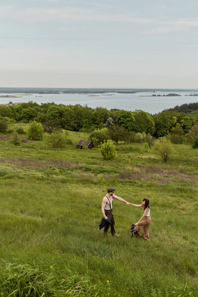 Вид позитивной и стильной пары в винтажных нарядах, держащейся за руки и стоящей на холме с сельским пейзажем на заднем плане, стильная пара, наслаждающаяся деревенской жизнью