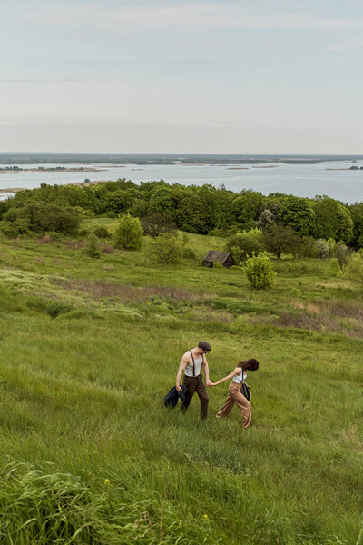 Брюнетка в стильном винтажном наряде держит за руку бойфренда в солнечных очках и подтяжках и ходит по травяному холму с природой на заднем плане, стильная пара наслаждается загородной жизнью