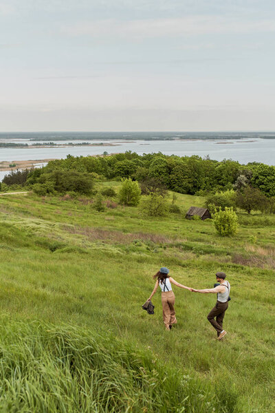 Модная романтическая пара в винтажных нарядах и кепках газетчиков, держащихся за руки и идущих по травяному холму с природой на заднем плане, стильная пара, наслаждающаяся загородной жизнью