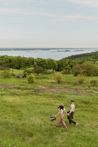 Модный мужчина в солнцезащитных очках и винтажном наряде, держащий за руку подругу в кепке ньюсмейкера и гуляющий вместе на фоне природы, стильная пара, наслаждающаяся деревенской жизнью