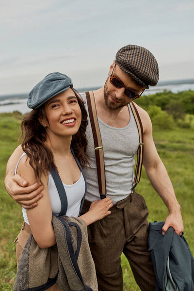 Модный мужчина в солнцезащитных очках и подтяжках обнимает весёлую брюнетку и держит куртку во время прогулки по размытому травянистому полю, модная пара на свежем воздухе