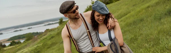 stock image Smiling and fashionable man in sunglasses and suspenders hugging brunette girlfriend in newsboy cap and walking together on grassy field at background, trendy couple in the rustic outdoors, banner 