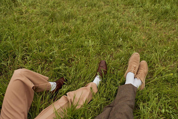 Top view of legs of romantic couple in retro-styled pants and shoes sitting next to each other on grassy meadow, stylish partners in rural escape, romantic getaway