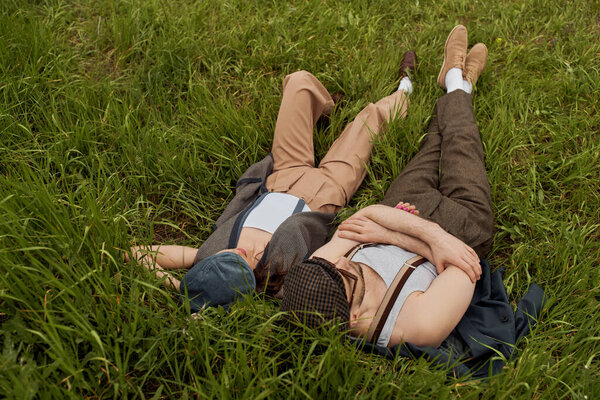 High angle view of fashionable romantic couple in newsboy caps and vintage outfits lying, spending time and relaxing together on grassy meadow, stylish partners in rural escape, romantic getaway