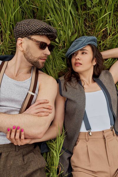 Top view of fashionable romantic couple in newsboy caps, suspenders and vintage outfits looking at each other while lying and relaxing on grassy field, stylish partners in rural escape