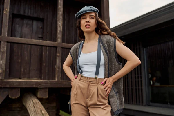 Stylish brunette woman in vintage outfit and newsboy cap posing in vest and suspenders while standing near rustic house in rural setting at summer, vintage-inspired clothing