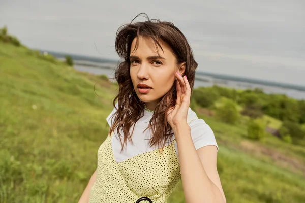 Portrait of stylish brunette woman in sundress touching face and looking at camera while standing with blurred scenic landscape and cloudy sky at background, summertime joy