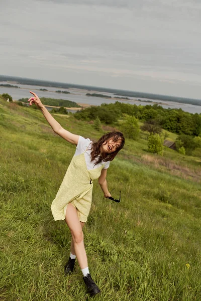 Stylish brunette woman in sundress and trendy boots holding sunglasses while having fun and standing on grass with scenic landscape at background at summer, summertime joy