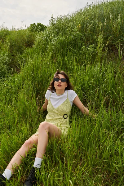 stock image Fashionable brunette woman in sunglasses and sundress spending time and relaxing on green meadow with grass, peaceful retreat and relaxing in nature concept, stylish woman