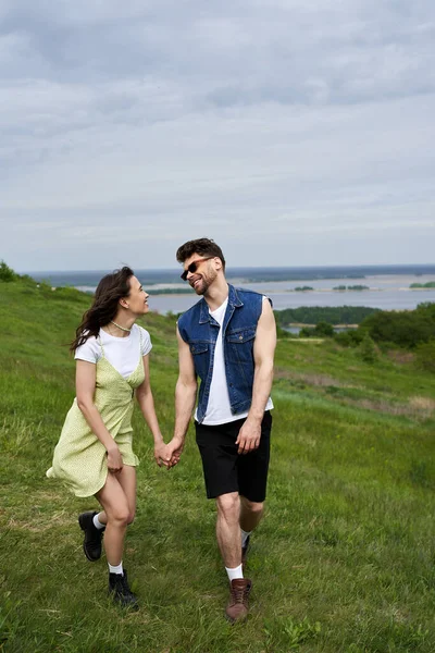 Positive and stylish brunette woman in sundress and boots holding hand and looking at boyfriend in sunglasses and walking together on grassy field, couple in love enjoying nature, tranquility