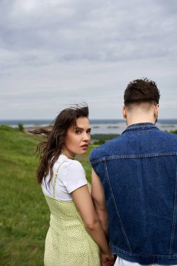Fashionable brunette woman in summer sundress looking at camera and holding hand of boyfriend in denim vest and standing on blurred green field, couple in love enjoying nature, tranquility clipart