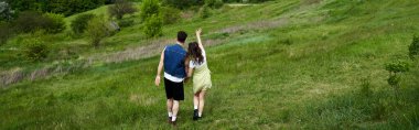 Back view of trendy romantic couple in summer outfits holding hands and standing on grassy hill with landscape at background, couple in love enjoying nature, banner, tranquility clipart