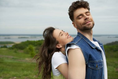 Cheerful brunette woman with closed eyes standing next to stylish bearded boyfriend in denim vest and spending time together with landscape at background, love story and countryside adventure clipart