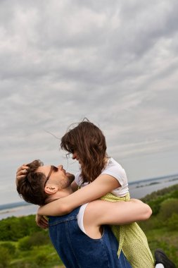 Gülümseyen, güneş gözlüklü, kot ceketli, esmer kız arkadaşına sarılıp kaldıran ve kırsal kesimde aşk hikayeleri, kırsal kesimde macera yaşayan bir adamın yan görüntüsü.