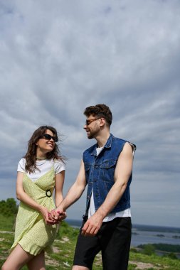 Gülümseyen esmer kadın güneş gözlüklü ve şık elbisesi olan yaz kıyafetleri ve kot yelek giyen sakallı erkek arkadaşıyla kırsal alanda gezinti tutkusu ve aşk konseptiyle zaman geçiriyor.