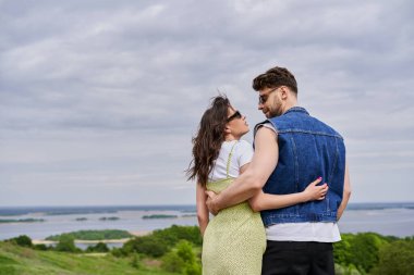 Güneş gözlüklü, yaz kıyafetleriyle birbirine sarılan ve manzara manzarası olan bulutlu arka planda gezintiler yapan romantik çiftin yan görüntüsü.