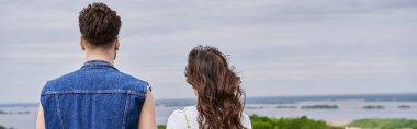 Back view of stylish brunette romantic couple in summer outfits standing with blurred cloudy sky and rural landscape at background, countryside retreat concept, banner clipart