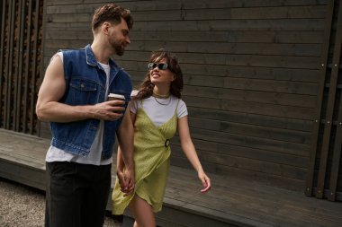 Smiling and stylish brunette woman in sunglasses and sundress holding hand of bearded boyfriend holding coffee to go while walking near wooden house in rural setting, outdoor enjoyment concept clipart