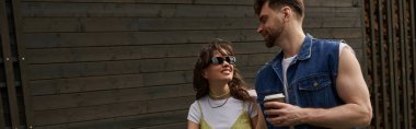 Smiling brunette woman in sunglasses and summer outfit looking at bearded boyfriend holding coffee to go while walking near wooden house at background, outdoor enjoyment concept, banner  clipart