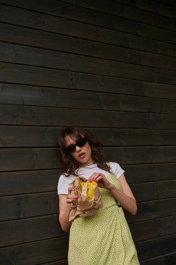 Trendy brunette woman in sunglasses and summer outfit holding fresh bun in craft package while standing near wooden house at background, summer vibes concept, tranquility clipart