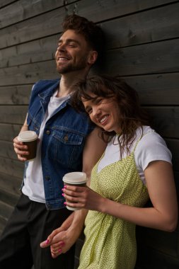 Cheerful and stylish brunette woman in summer outfit holding hand of bearded boyfriend in denim jacket and coffee to go and standing near wooden house, carefree moments concept clipart