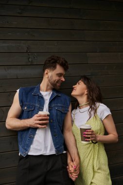 Cheerful and stylish romantic couple in summer outfits looking at each other and holding coffee to go while standing near wooden house in rural setting, carefree moments concept clipart