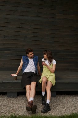 Full length of joyful brunette woman in boots and sundress holding coffee to go and talking to bearded boyfriend in denim vest while sitting near wooden house, serene ambiance concept clipart