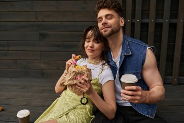 Positive bearded man in denim vest holding coffee to go and sitting near stylish brunette girlfriend in sundress holding tasty bun and wooden house at background, serene ambiance concept clipart