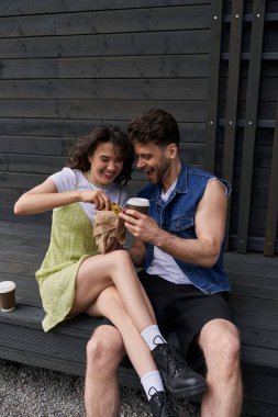 Positive brunette woman in boots and sundress holding fresh bun near stylish boyfriend in denim vest with coffee to go while sitting near wooden house at background, serene ambiance concept clipart