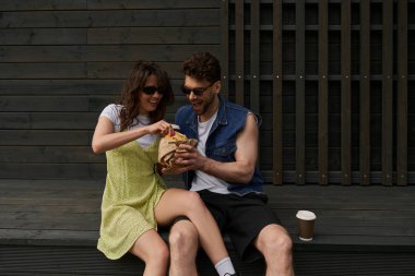 Neşeli ve modaya uygun, güneş gözlüklü romantik çift ve yaz kıyafetleri içinde taze çörek tutarken kahve yanına oturup arka planda ahşap ev, kırsal kesim keşfi konsepti