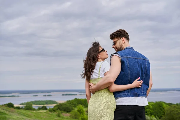 Side view of trendy romantic couple in sunglasses and summer outfits hugging and looking at each other with scenic landscape and cloudy sky at background, countryside leisurely stroll