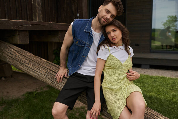 Smiling and stylish bearded man in denim vest hugging brunette girlfriend in sundress and looking at camera while sitting on wooden log in rural setting, countryside retreat concept